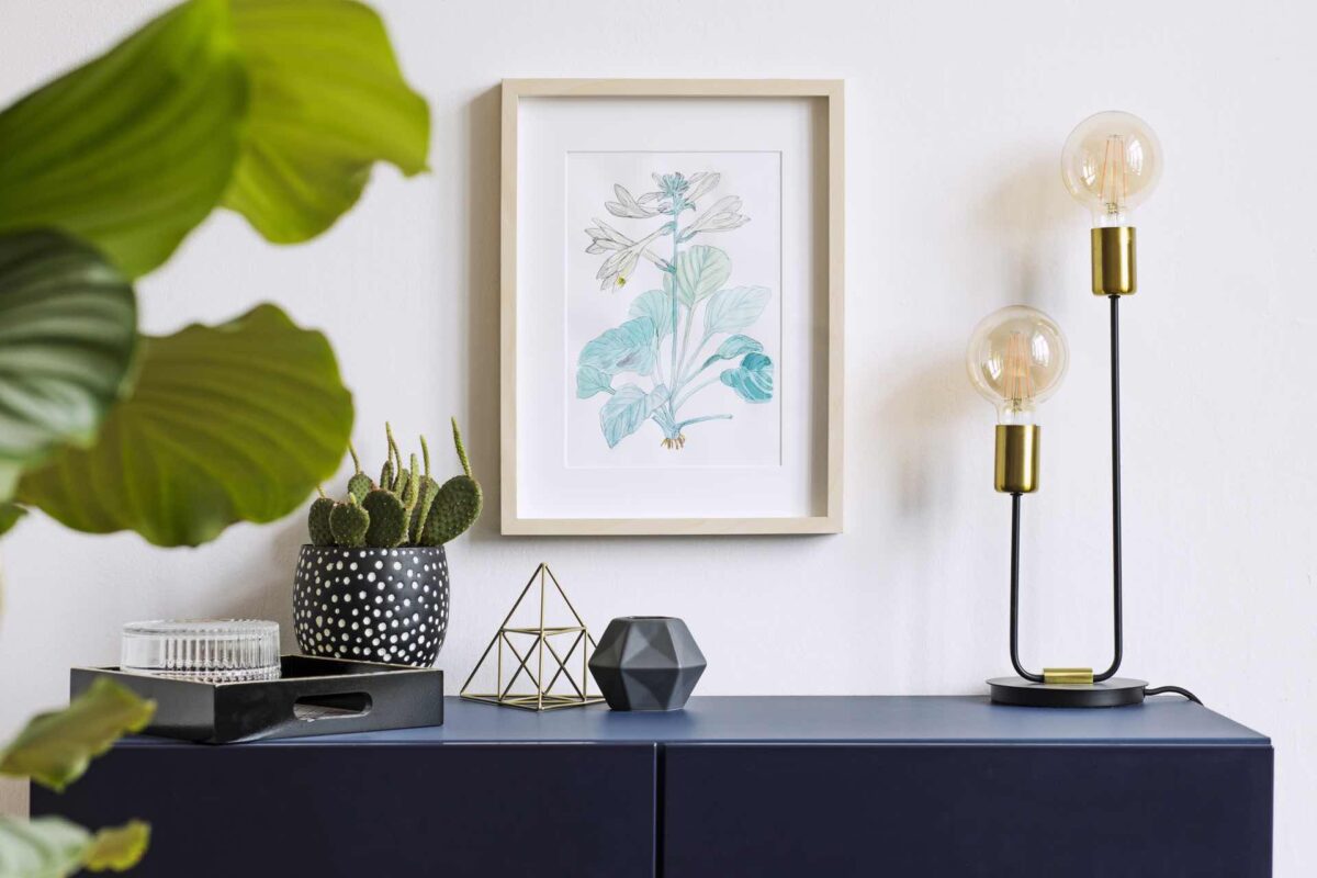 White interior of floral poster mock up with vertical wooden frame, table lamp, , cacti, gold pyramid and tropical flowers on the white wall background. Concept with navy blue shelf.
